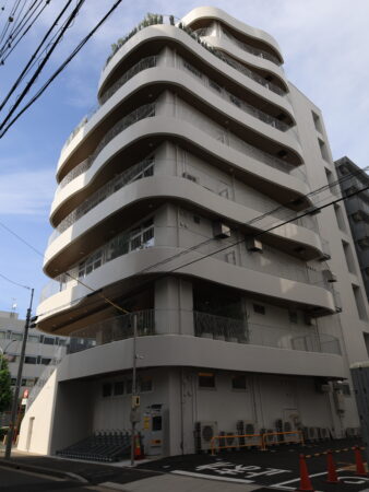 デザインビルのバルコニーを真下からダイナミックに見上げたカット。何層にも重なる有機的な曲線壁、温かみのある木目調の軒裏、繊細な縦格子手すりのディテールが強調されている。