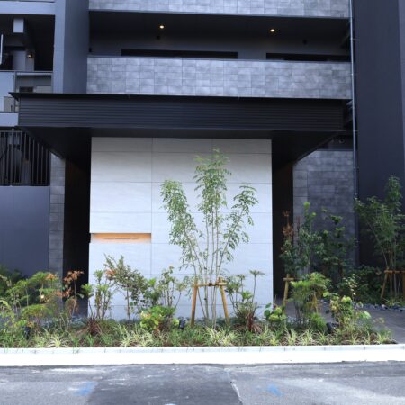 エントランス庇の正面全景。黒のアルミ幕板が、白いタイル壁と青々とした植栽を引き立てます。